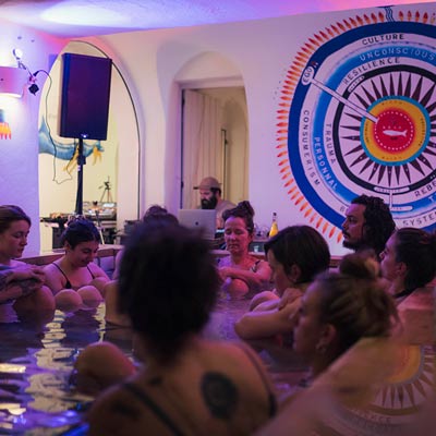 Personen sitzen in einem Holzbad in einem Raum in Thermalwasser und geniessen die musik die ein bärtiger Dj im Hintergrund abspielt.